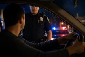 Police officer speaking with a driver through an open car window during a nighttime traffic stop, with red and blue patrol lights visible in the background.